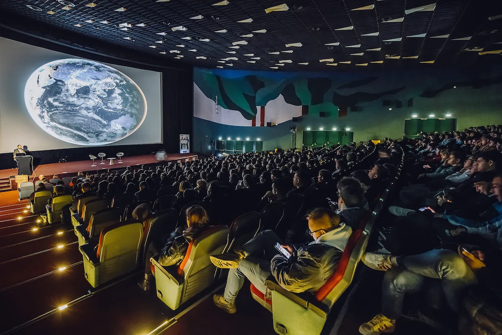 Sala kinowa wypełniona publicznością, wszyscy zwróceni w stronę dużego ekranu z wyświetlonym obrazem Ziemi. Na scenie osoba przemawiająca, a wnętrze oświetlone delikatnym światłem bocznym i dekoracyjne malowidła na ścianie.
