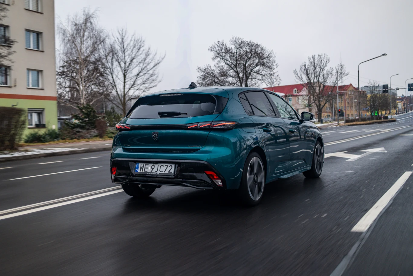 Nowoczesny zielony hatchback jadący po pustej miejskiej ulicy, otoczony niskimi budynkami mieszkalnymi i drzewami bez liści, przy pochmurnej pogodzie.