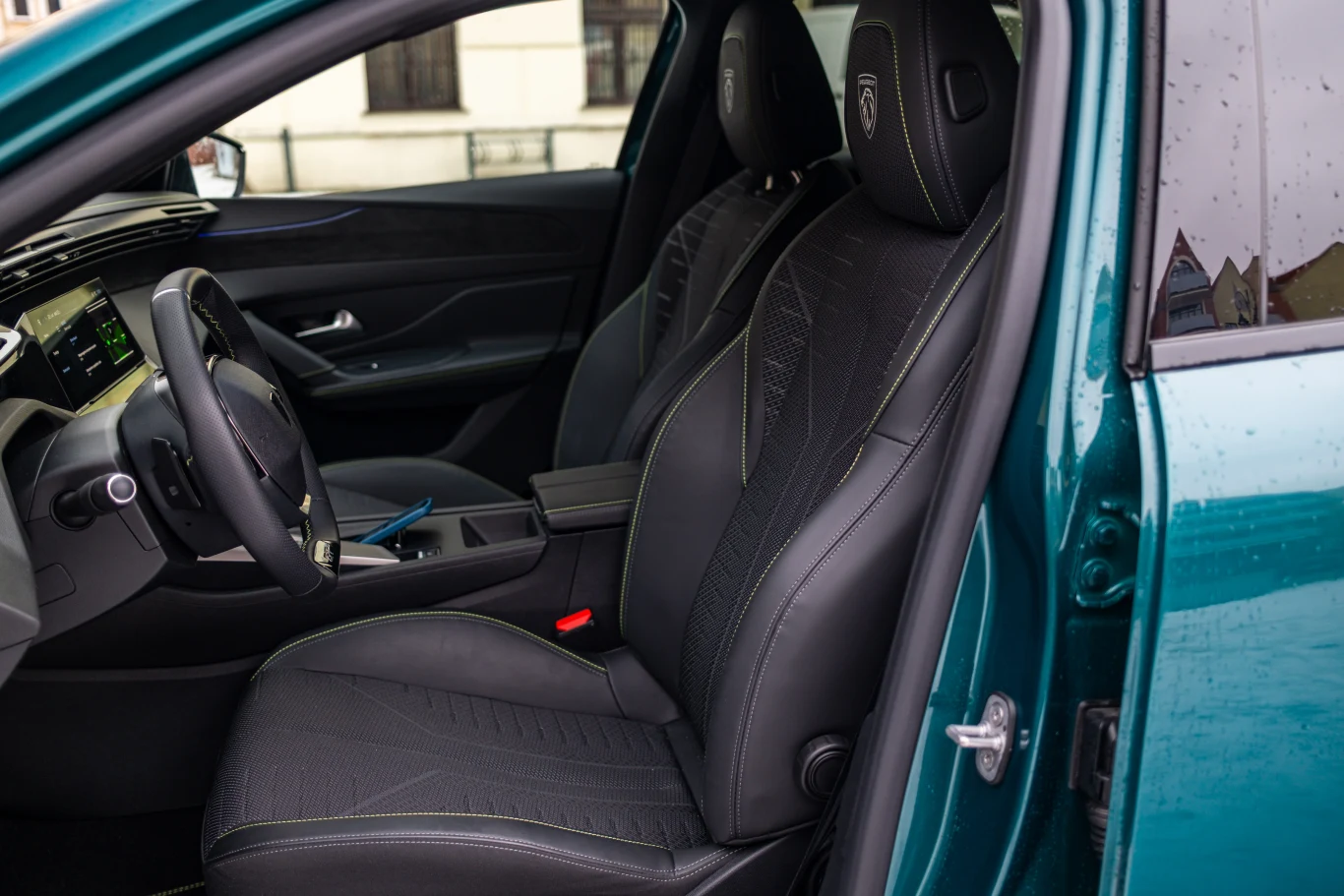 Wnętrze nowoczesnego samochodu z widocznym przednim siedzeniem kierowcy i pasażera, czarną tapicerką ze szwami oraz fragmentem deski rozdzielczej z wyświetlaczem i kierownicą.