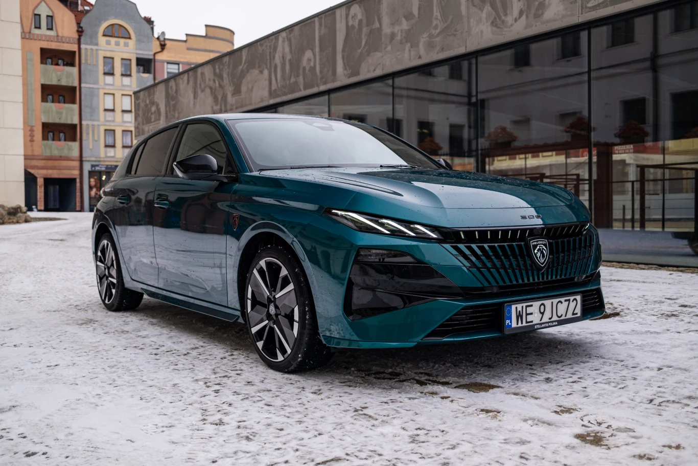 Nowoczesny, zielony hatchback marki Peugeot zaparkowany na ośnieżonej ulicy przy nowoczesnych i zabytkowych budynkach, z widoczną rejestracją i charakterystycznym logo na masce.