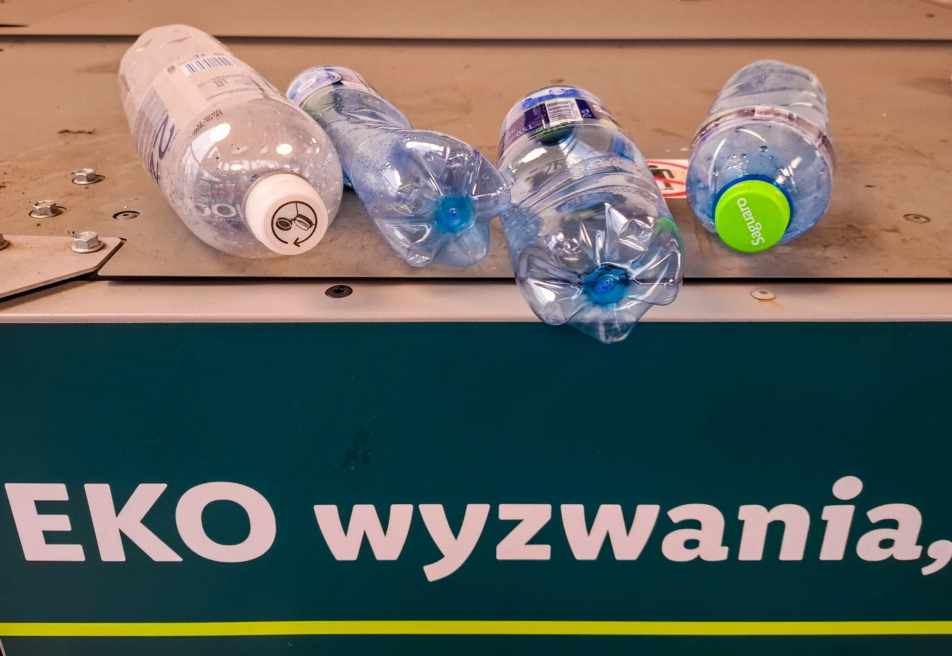Mimo że butelki po mleku, kefirach czy jogurtach pitnych są wykonane z tworzyw sztucznych są wykluczone z systemu kaucyjnego. Cztery zgniecione plastikowe butelki po wodzie mineralnej ułożone na zielonej powierzchni z napisem EKO wyzwania.