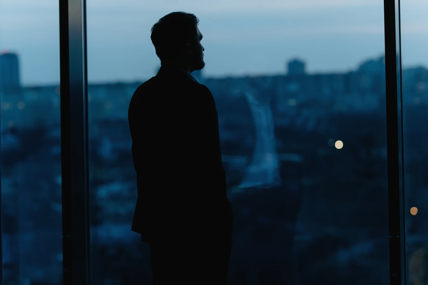 Sylwetka mężczyzny stojącego przy dużym oknie z widokiem na miasto po zmroku; postać jest ukazana w cieniu