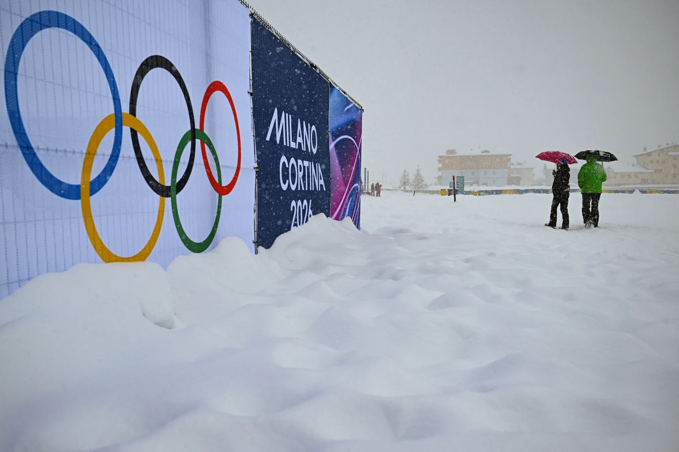 Zimowe igrzyska olimpijskie Duża ilość śniegu pokrywająca teren, logo igrzysk olimpijskich oraz napis Milano Cortina 2026 na banerze, dwie osoby stojące pod parasolami z dala od głównej sceny wydarzeń.