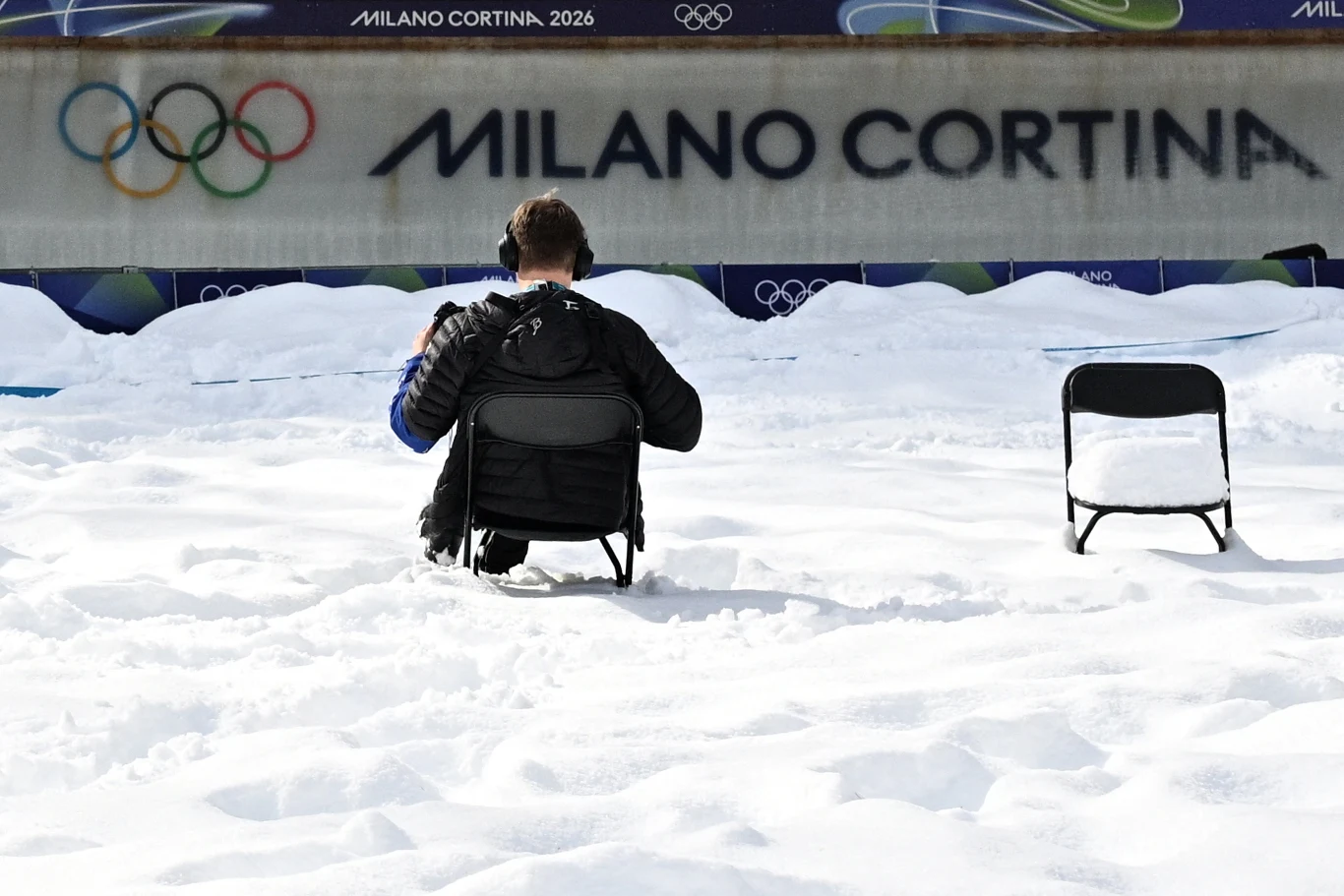 Tor lodowy w Cortinie d'Ampezzo Osoba siedząca na krześle na zaśnieżonym terenie, naprzeciw ściany z dużym napisem Milano Cortina oraz olimpijskimi kołami, obok puste krzesło.
