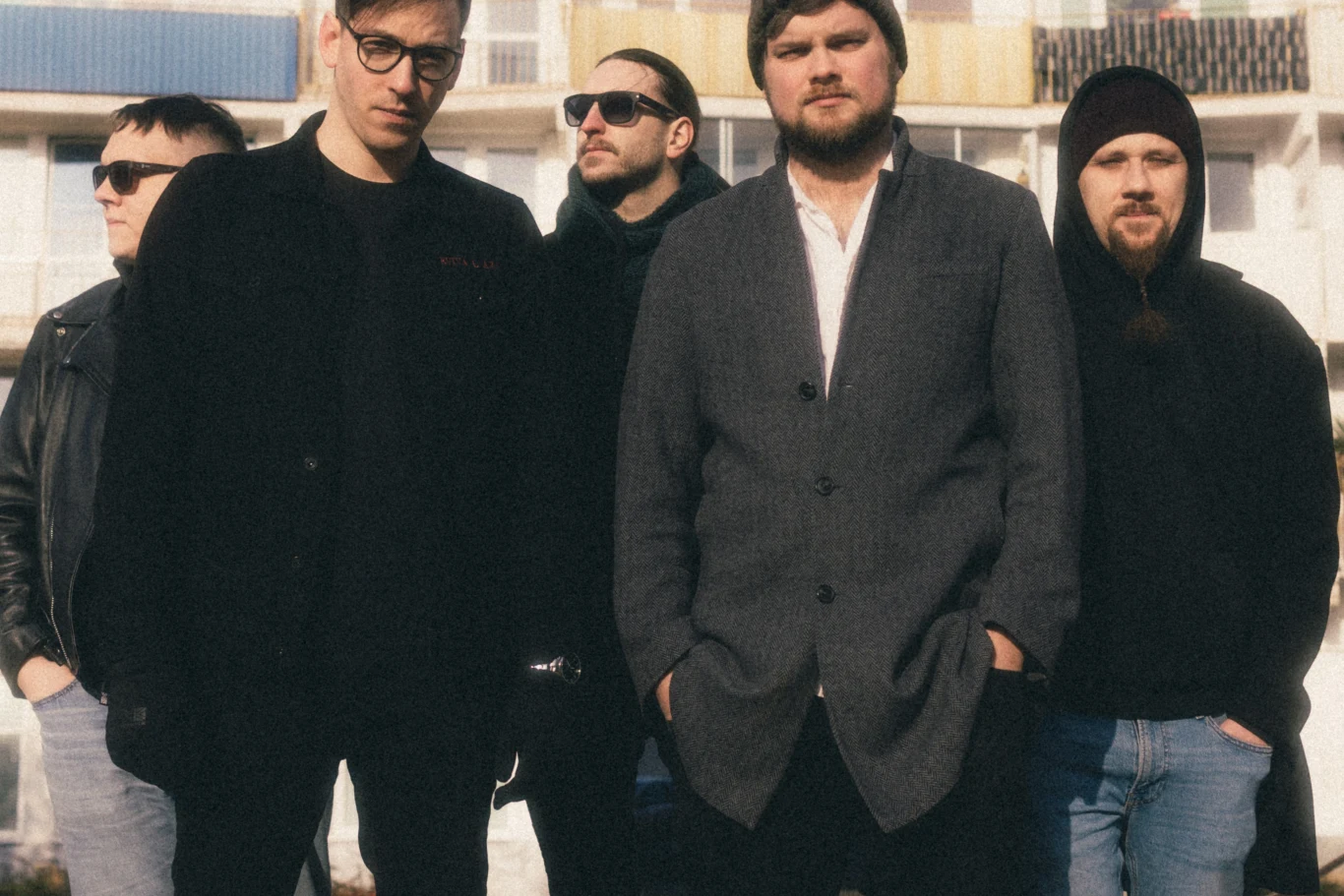 Zespół kast. Grupa pięciu mężczyzn w różnym wieku stoi razem na zewnątrz, na tle wielopiętrowego budynku mieszkalnego. Część osób ma założone okulary przeciwsłoneczne, jeden z mężczyzn jest ubrany w elegancki płaszcz, a reszta w bardziej codzienne stroje.