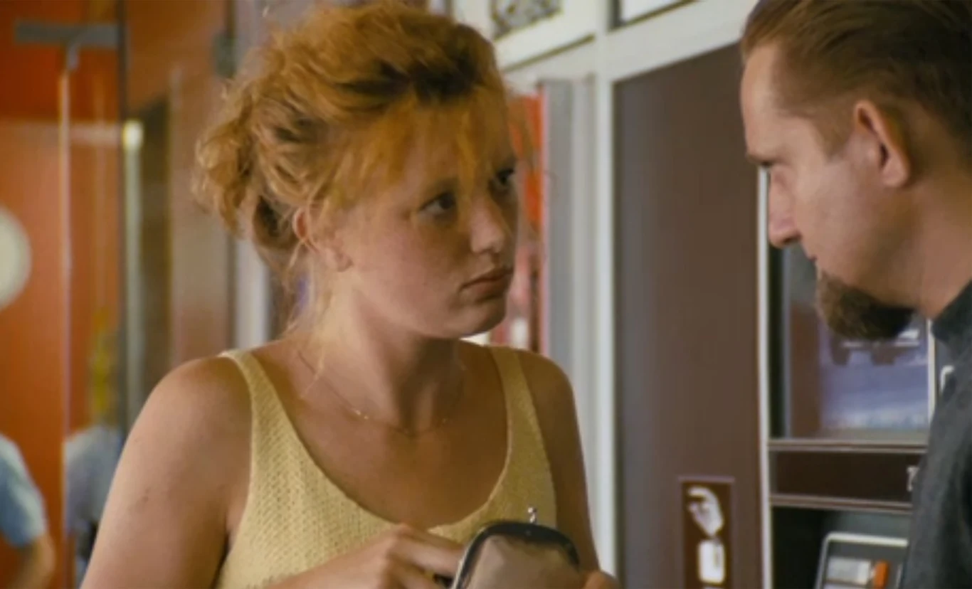Młoda kobieta o rudych włosach w żółtej bluzce spogląda poważnie na mężczyznę stojącego naprzeciwko niej, w ręku trzyma portfel, w tle elementy wnętrza lokalu.