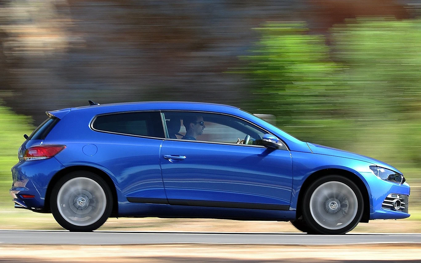 Niebieski samochód osobowy w ruchu na tle rozmytego krajobrazu, widoczny z boku, jadący po asfaltowej drodze.