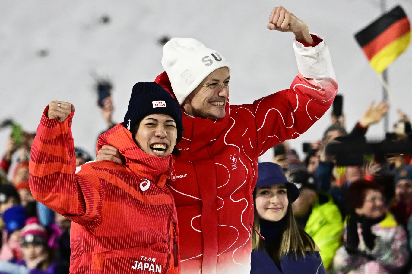 Dwóch sportowców ubranych w charakterystyczne stroje reprezentacyjne świętuje sukces, unosząc zaciśnięte pięści w geście triumfu. W tle tłum kibiców, widoczne narodowe flagi, w tym flaga Niemiec, oraz zimowa, śnieżna sceneria.