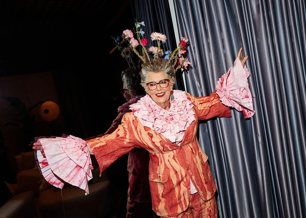 Prue Leith podczas London Fashion Week w 2026 r. Osoba w ekstrawaganckim stroju składającym się z kolorowego garnituru i dużych falban na rękawach oraz kołnierzu, z charakterystyczną fryzurą ozdobioną kwiatami, pozuje radośnie na tle zasłon.