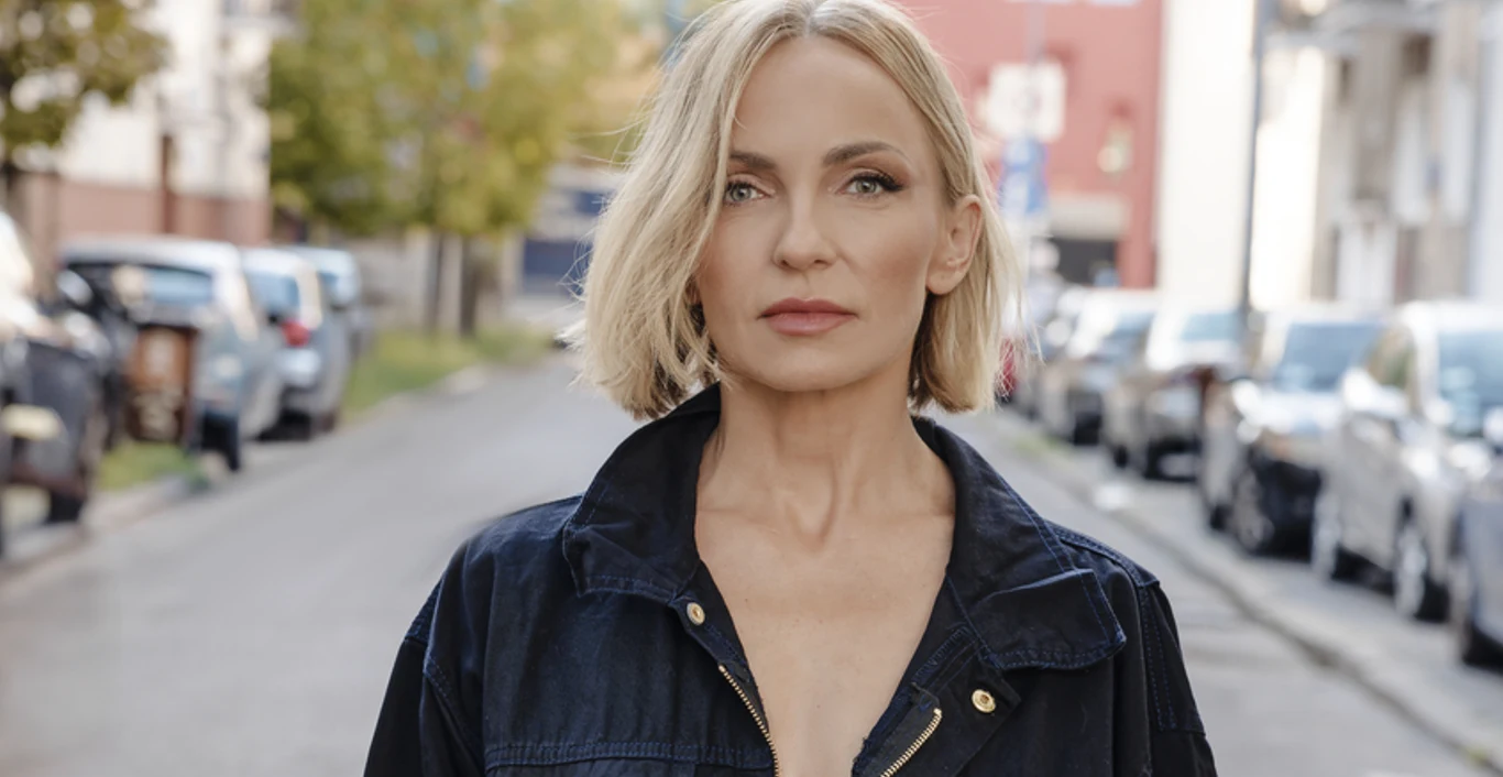 Ania Wyszkoni zaprasza do świata jej nowego albumu Kobieta o blond włosach i krótkiej fryzurze stoi na ulicy wśród zaparkowanych samochodów, ubrana w czarną kurtkę z dużym dekoltem, patrząca prosto w obiektyw.