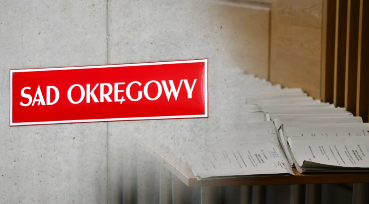 Wraca sprawa Roberta J. Czerwona tablica z napisem Sąd Okręgowy obok rzędu uporządkowanych akt sądowych leżących na regale, tło neutralne, wnętrze instytucji sądowej.