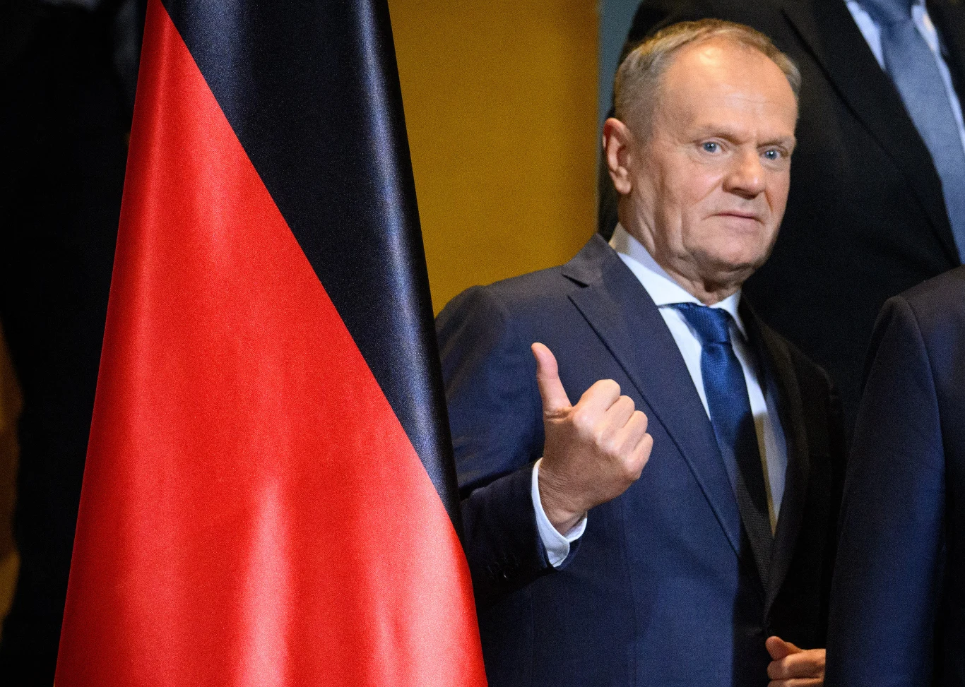 Mężczyzna w garniturze stoi obok dużej flagi w kolorach czarnym, czerwonym i żółtym, unosząc kciuk w geście aprobaty, patrzy bezpośrednio w stronę aparatu.