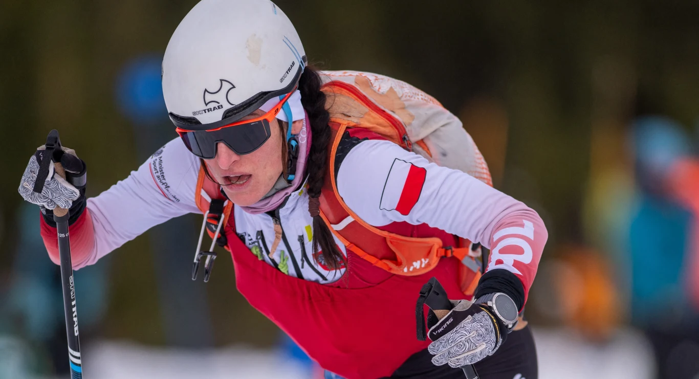 Zawodniczka w zimowej odzieży sportowej z polską flagą na ramieniu, w kasku i goglach, biorąca udział w zawodach narciarskich w otoczeniu śniegu i drzew.