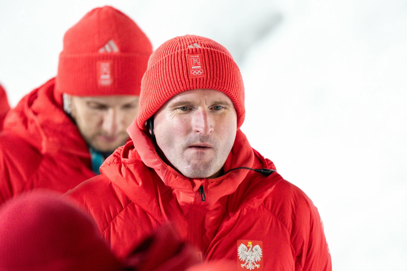 Maciej Maciusiak Mężczyzna ubrany w czerwoną kurtkę sportową z orłem oraz czerwoną czapkę z godłem Polski i napisem „POL”, stojący na śniegu, w tle inna osoba ubrana podobnie.