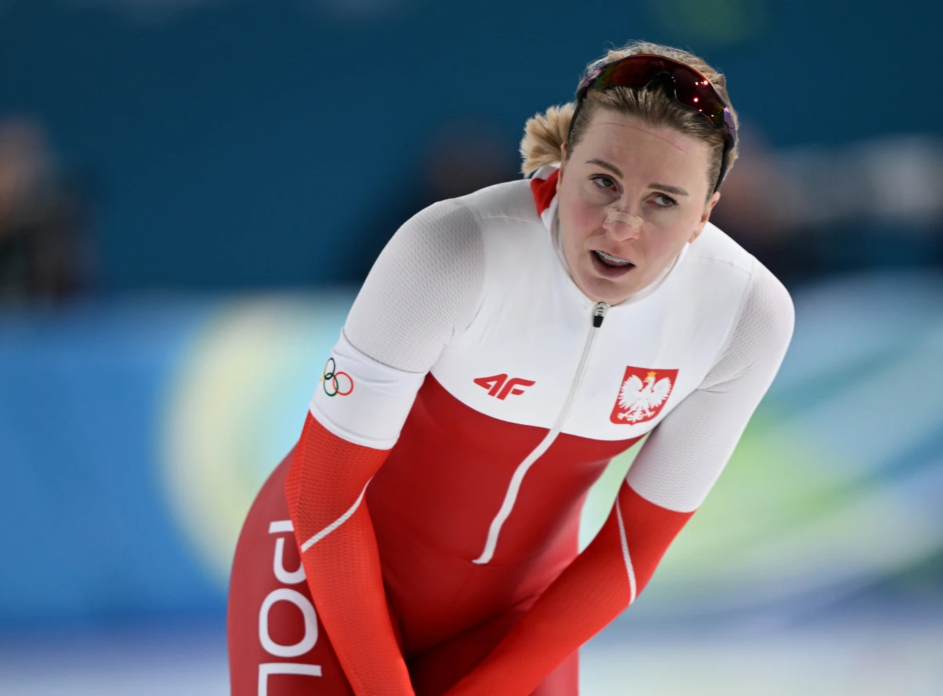 Natalia Czerwonka Zawodniczka w czerwono-białym stroju z napisem 'POLSKA' oraz godłem narodowym, lekko pochylona do przodu na lodowisku, w tle rozmazane niebieskie tło.