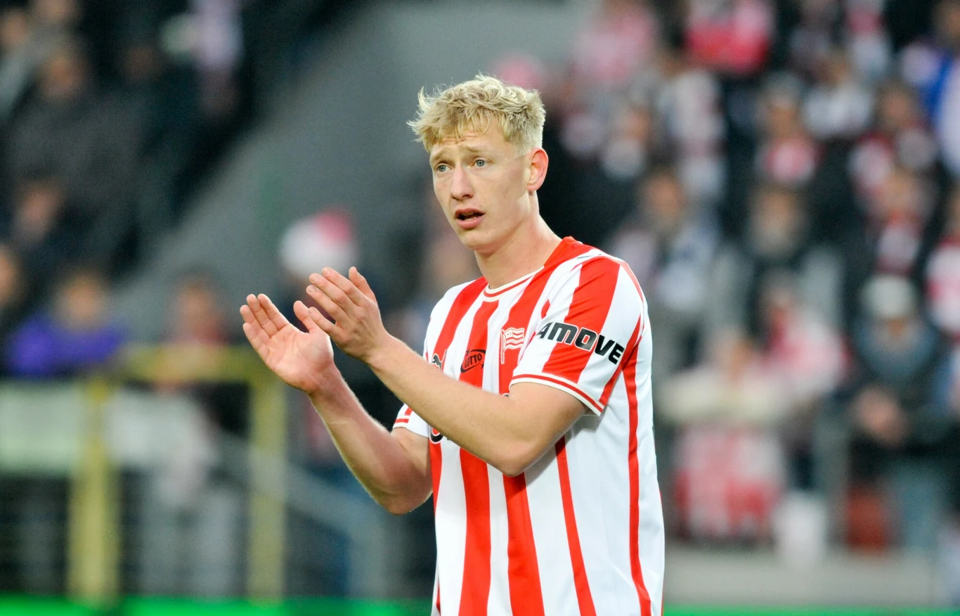 Oskar Wójcik Młody mężczyzna w czerwono-białej piłkarskiej koszulce znajduje się na stadionie, prawdopodobnie podczas meczu, bije brawo, a w tle rozmazane sylwetki kibiców.