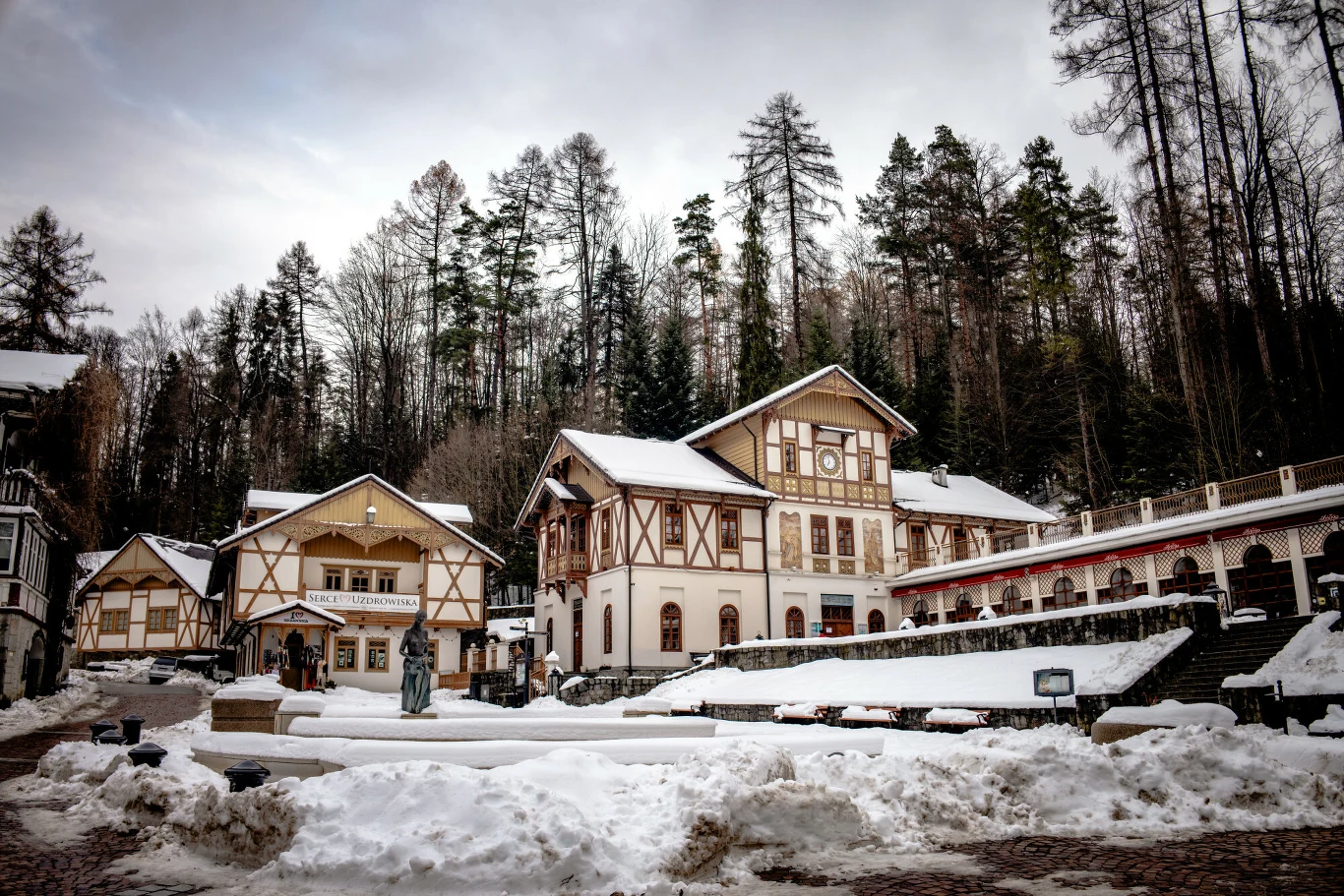 Pijalnia wód w Szczawnicy Kompleks budynków w stylu alpejskim otoczonych zimowym krajobrazem, pokryte śniegiem dachy i tereny wokół, w tle las iglasty na zboczu góry.
