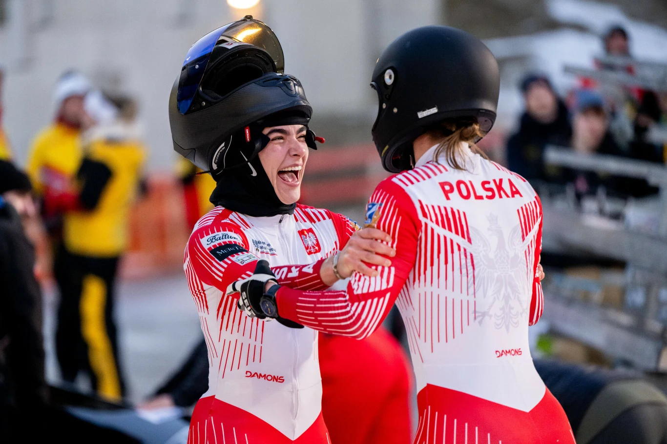Dwie zawodniczki polskiej drużyny w strojach sportowych i kaskach cieszą się i gratulują sobie po zakończeniu wyścigu, w tle widoczni inni uczestnicy imprezy sportowej.