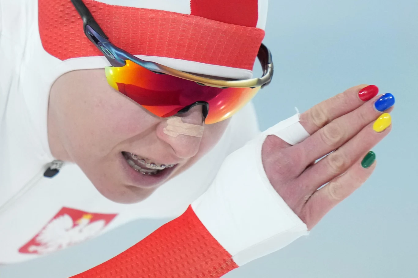 Łyżwiarka szybka w biało-czerwonym stroju z orłem na piersi, w sportowych okularach, podczas dynamicznego ruchu, z kolorowymi paznokciami podkreślającymi olimpijskiego ducha.