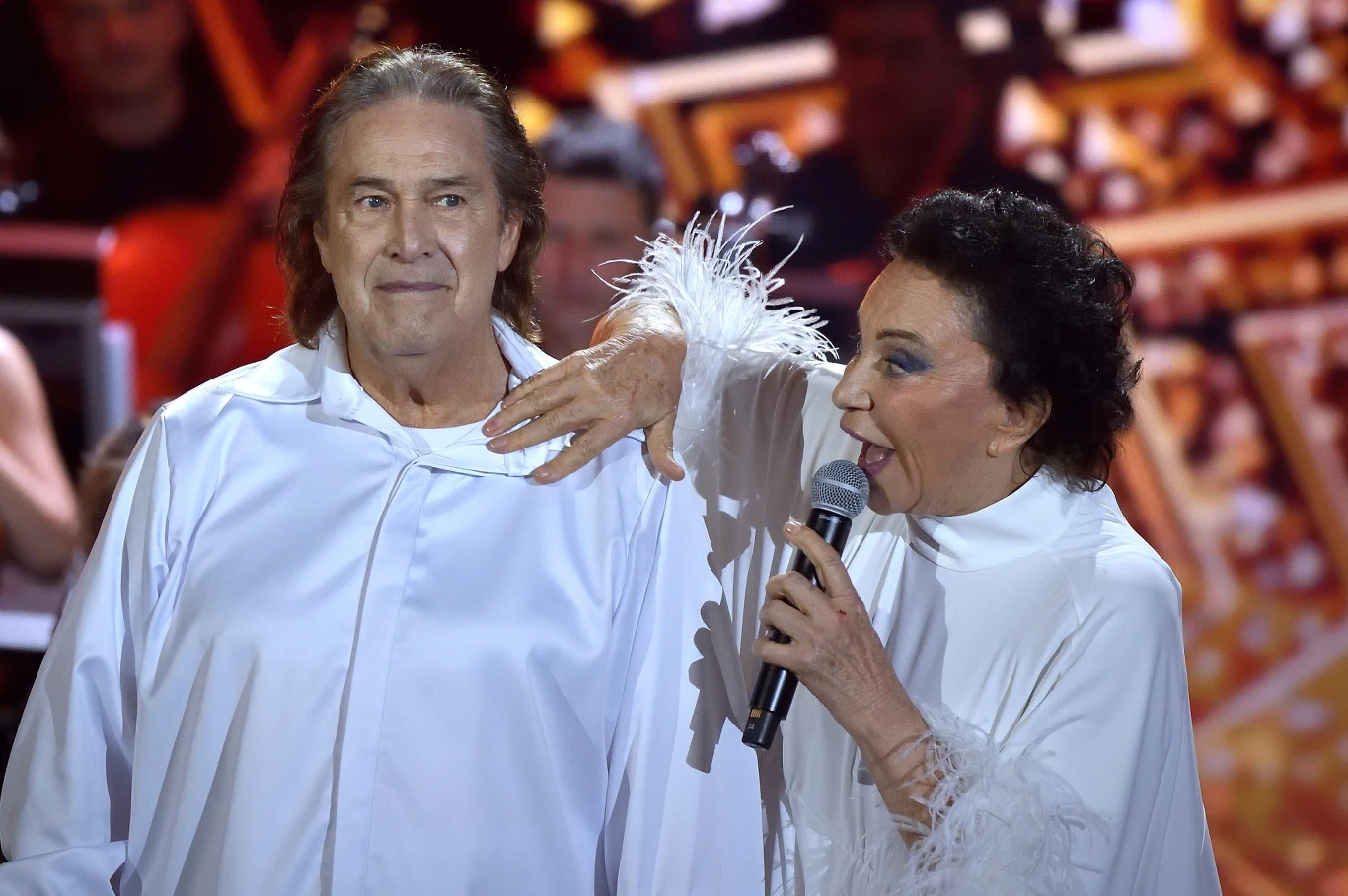 Dwójka artystów w białych strojach sceniczych, stojąca na tle rozświetlonej sceny, jedna osoba trzyma mikrofon i gestykuluje w kierunku drugiej osoby.