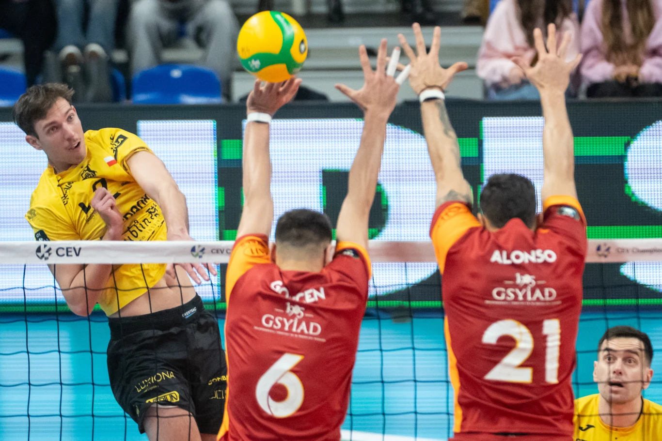 Siatkarz w żółtym stroju atakuje piłkę przez siatkę, naprzeciwko dwaj zawodnicy w czerwonych koszulkach próbują ją zablokować, w tle widoczna publiczność.