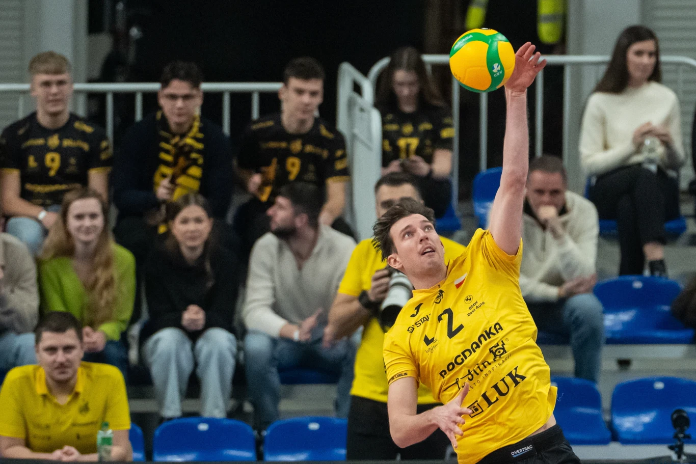 Siatkarz w żółtej koszulce wykonuje zagrywkę podczas meczu, w tle widać kibiców siedzących na trybunach, część z nich ubrana w klubowe barwy.