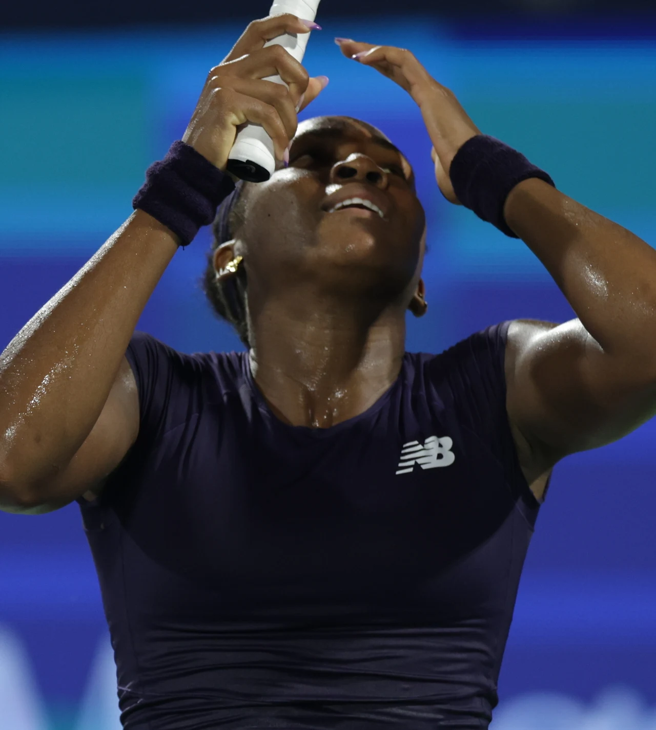 Zawodniczka tenisowa w ciemnym stroju sportowym, trzymająca rakietę i dotykająca czoła, na jej skórze widoczne są krople potu, tło utrzymane w błękitnych kolorach rozmazanych przez ruch sportowy.