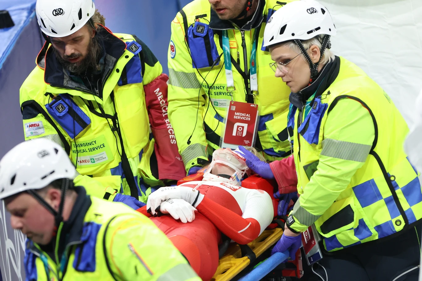 Ratownik medyczny oraz trzech asystentów transportuje sportowca ubranego w czerwony kombinezon na noszach. Sportowiec ma założony kołnierz ortopedyczny oraz maskę tlenową, a personel medyczny jest ubrany w jaskrawe stroje ochronne i kaski.