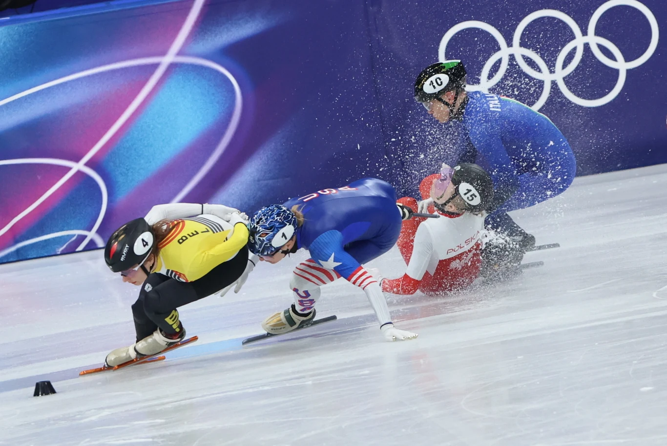 Zawodniczki w trakcie wyścigu łyżwiarskiego na lodzie, jedna z nich upada, podczas gdy reszta kontynuuje jazdę, widoczny śnieg lodowy rozpryskuje się wokół, w tle symbole olimpijskie.