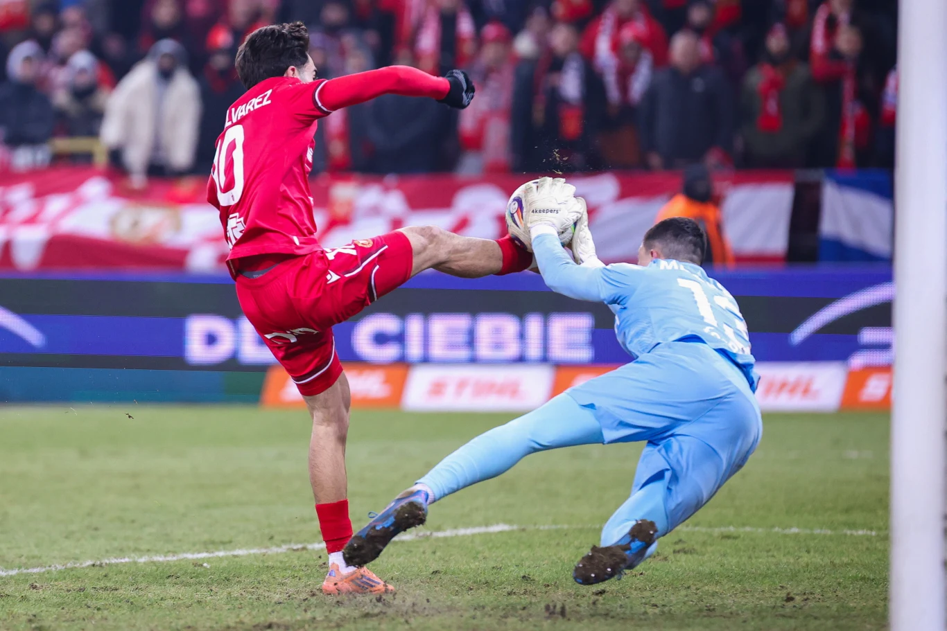 Piłkarz w czerwonym stroju oddaje strzał na bramkę, podczas gdy bramkarz w niebieskim stroju próbuje obronić piłkę w dynamicznej akcji na boisku futbolowym; w tle widoczni kibice z flagami.