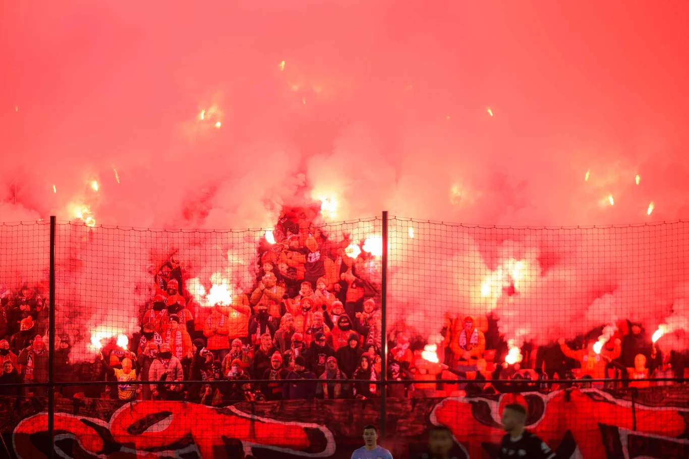 Grupa kibiców na stadionie unosi w górę race świetlne, wokół rozświetlone czerwonym światłem, nad tłumem unosi się gęsty dym, w tle widoczne siatki zabezpieczające i fragmenty graffiti.