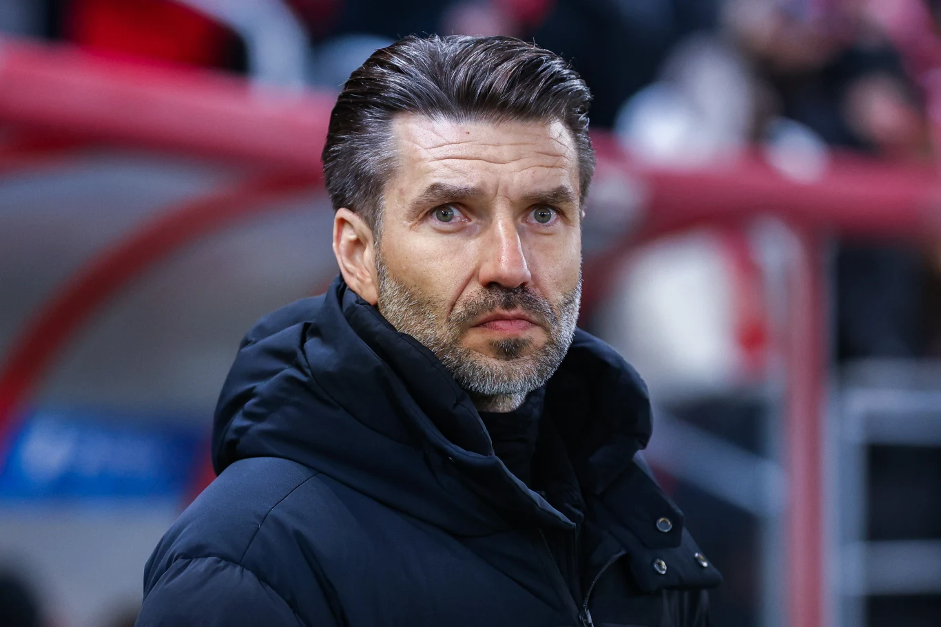 Mężczyzna o siwiejących włosach i brodzie stoi na tle trybun, ubrany w ciemną kurtkę zimową, patrzy poważnie przed siebie.