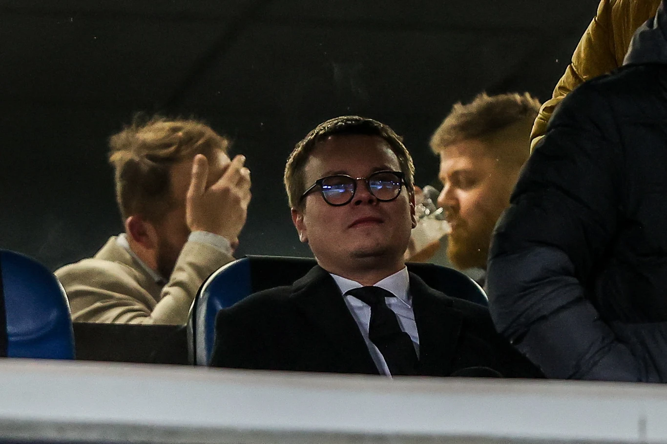 Prezes Wisły Jarosław Królewski po meczu spieszył się na spotkanie z Wojciechem Kwietniem (właściciel Wieczystej) oraz Bogusławem Cupiałem (były właściciel Wisły) Mężczyzna w garniturze i okularach siedzi na trybunach, w tle dwóch innych mężczyzn, z których jeden pije napój z plastikowego kubka, a drugi poprawia włosy.