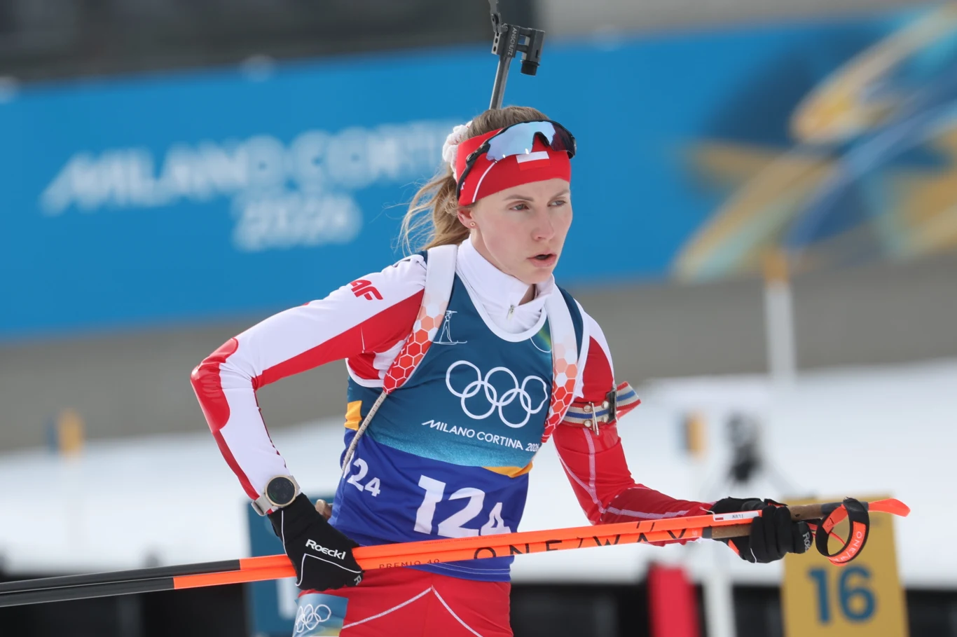 Zawodniczka w stroju reprezentacyjnym podczas zawodów biathlonowych na zimowych igrzyskach olimpijskich, z karabinem na plecach, w otoczeniu zimowej scenerii stadionu sportowego.