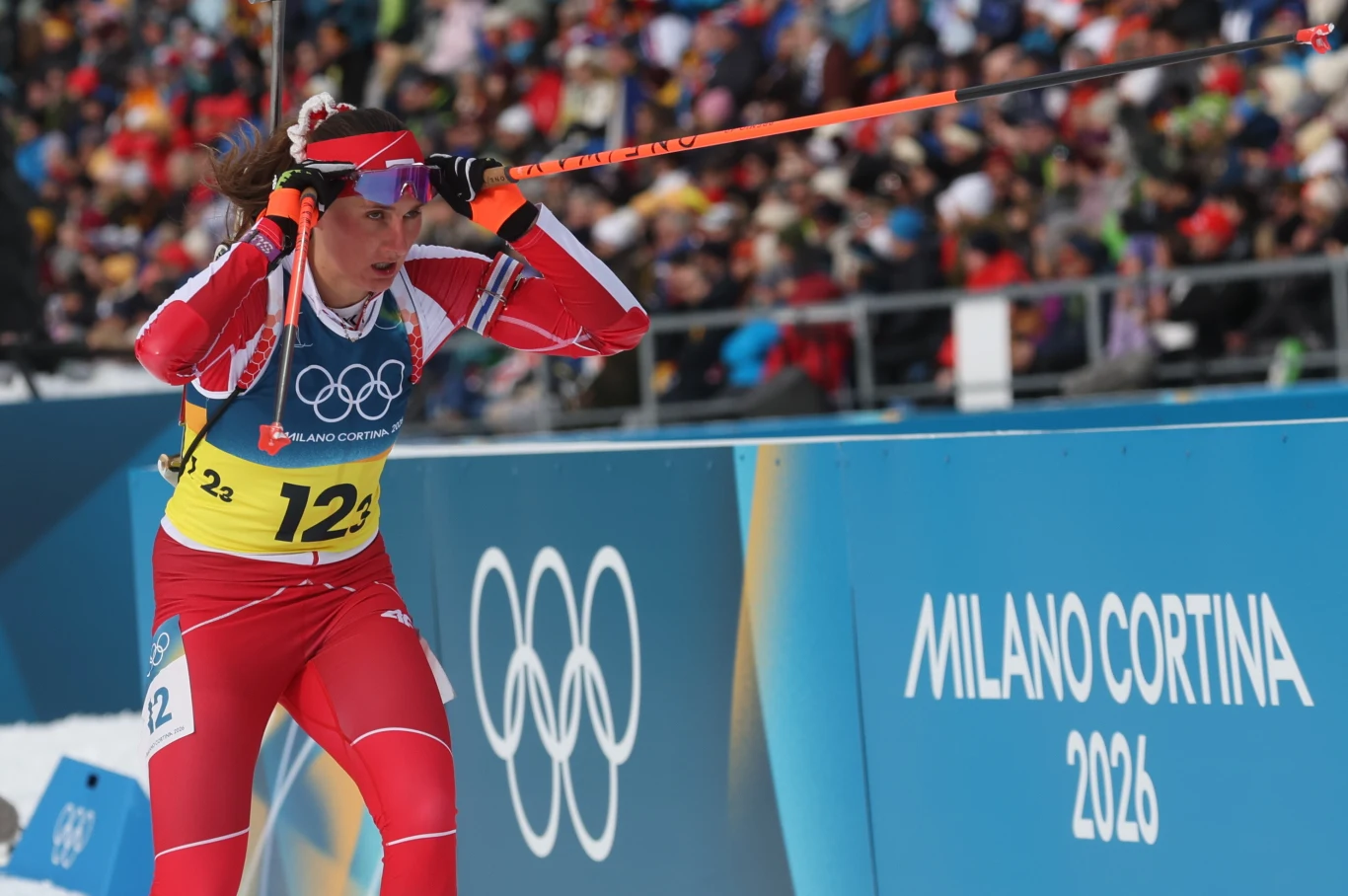 Zawodniczka biegnąca na nartach w trakcie zawodów olimpijskich, ubrana w czerwony kombinezon startowy oraz żółty numer startowy 123, trzymająca kijki narciarskie, w tle licznie zgromadzona publiczność i duży niebieski baner z napisem Milano Cortina 202...