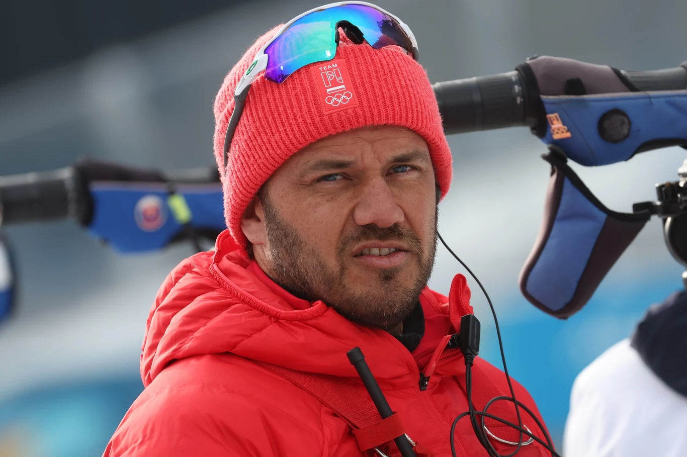 Mężczyzna w czerwonej kurtce i czapce z logo olimpijskim, z niebieskimi okularami narciarskimi na głowie, stoi na tle sprzętu sportowego używanego do biatlonu.