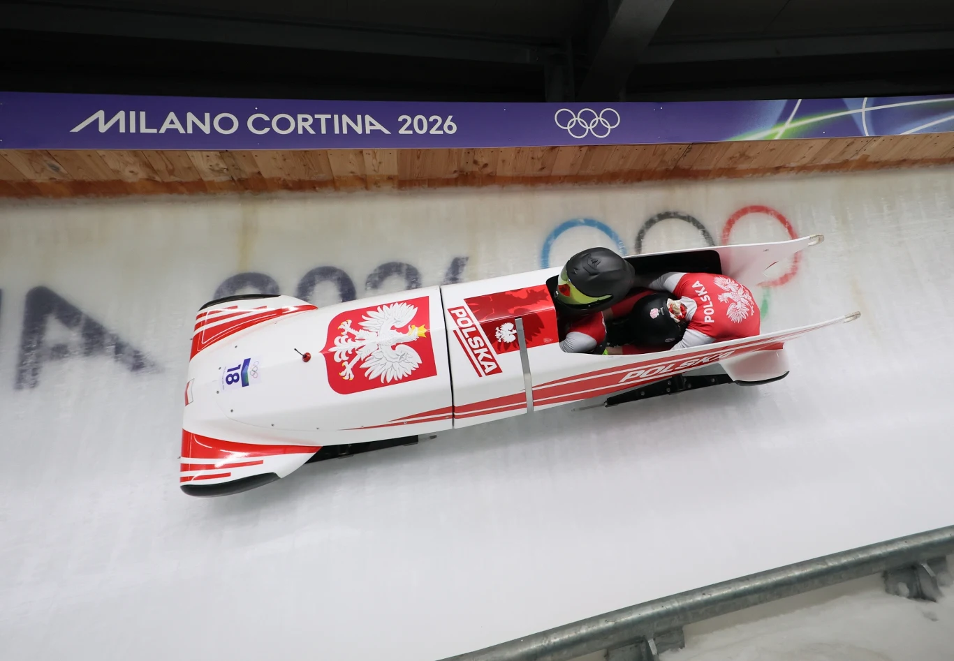 Polska załoga bobslejowa zjeżdża lodowym torem bobslejowym podczas przygotowań do Zimowych Igrzysk Olimpijskich Milano Cortina 2026. Bobslej zdobiony biało-czerwonymi barwami i godłem Polski. Załoga w kaskach, skupiona na sportowej rywalizacji.