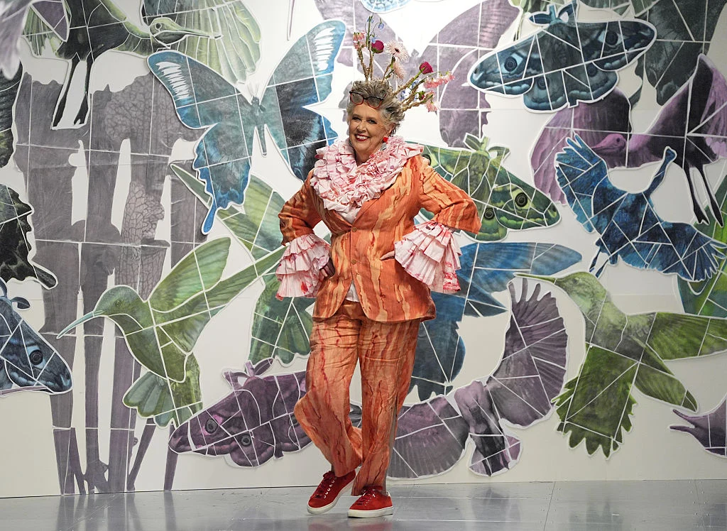 Prue Leith w garniturze z innowacyjnej tkaniny Kobieta w kolorowym stroju teatralnym z bogato zdobioną koroną na głowie i szerokimi rękawami pozuje na tle ściany ozdobionej dużymi grafikami ptaków i motyli.
