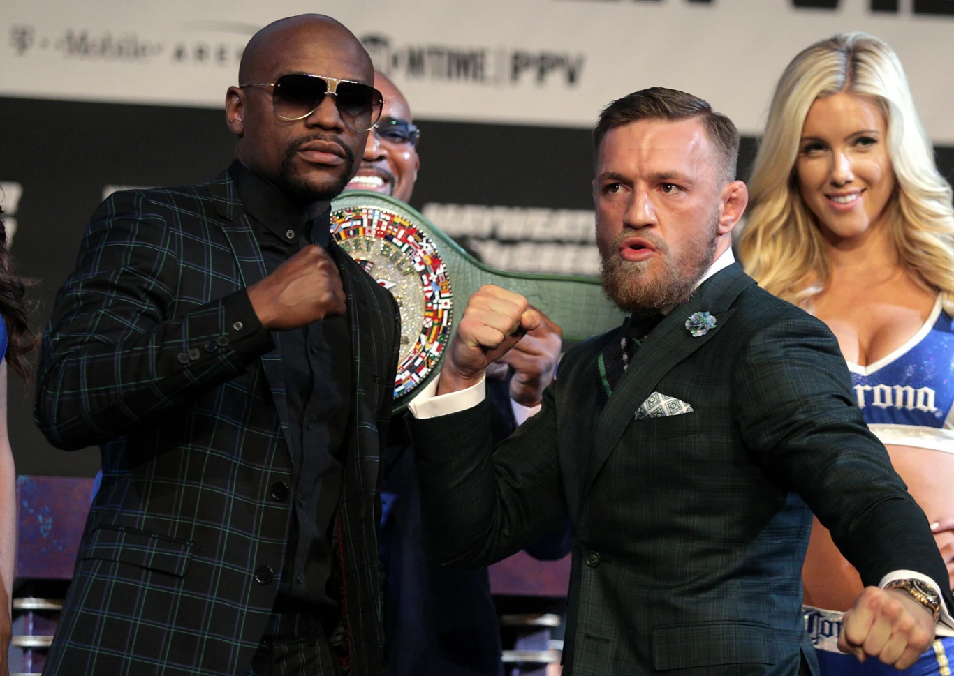 Floyd Mayweather i Conor McGregor, 23.08.2017 r. Dwaj mężczyźni w eleganckich garniturach stoją naprzeciw siebie z zaciśniętymi pięściami w bojowej pozie, w tle widoczny pas mistrzowski oraz uśmiechnięta kobieta w stroju z napisem Corona.