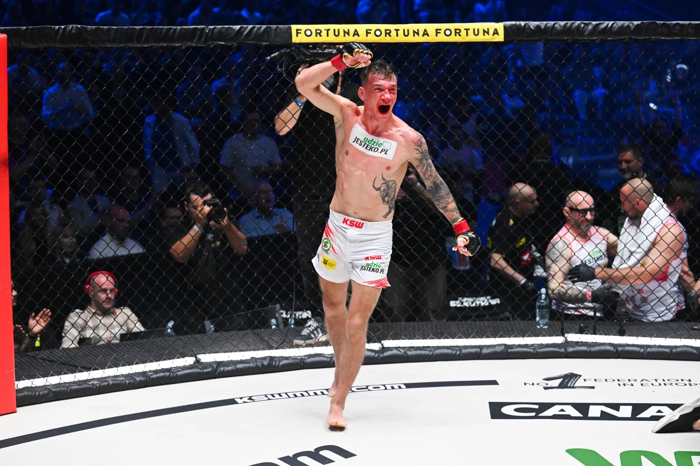 Zawodnik MMA unosi rękę w geście triumfu w oktagonie, z oznakami krwi na twarzy i tułowiu, otoczony przez sędziów, rywali oraz publiczność obserwującą wydarzenie sportowe.