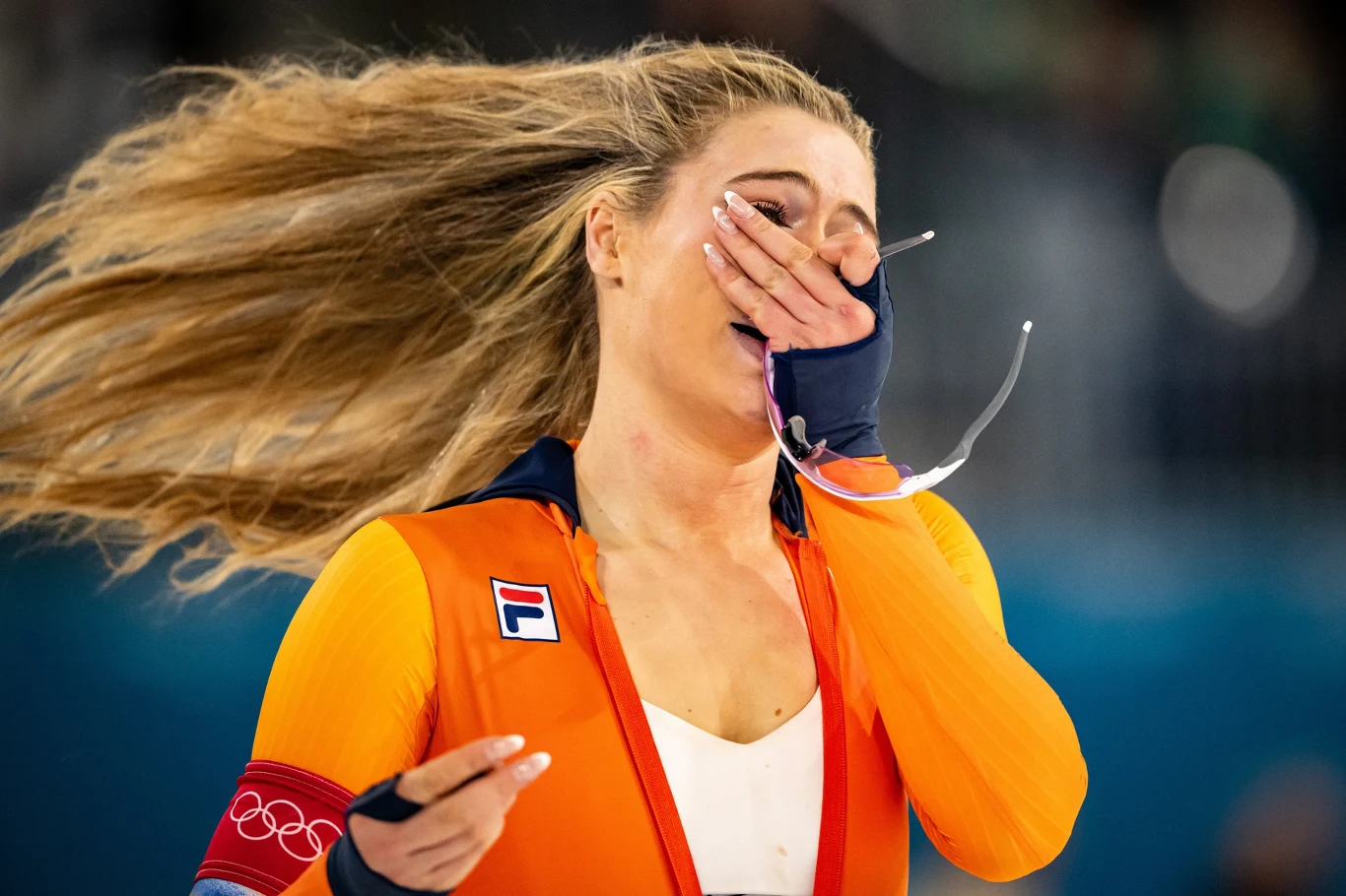 Sportsmenka w pomarańczowym stroju olimpijskim z długimi, rozwianymi włosami, z ręką przy twarzy, wyrażając silne emocje po zakończeniu zawodów, widoczne łzy wzruszenia na twarzy.