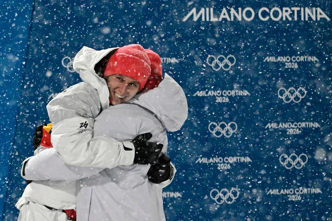 Dwoje sportowców ubranych w białe kurtki przytula się z radością podczas śnieżycy na tle niebieskiej ścianki z logo igrzysk olimpijskich Milano Cortina 2026.