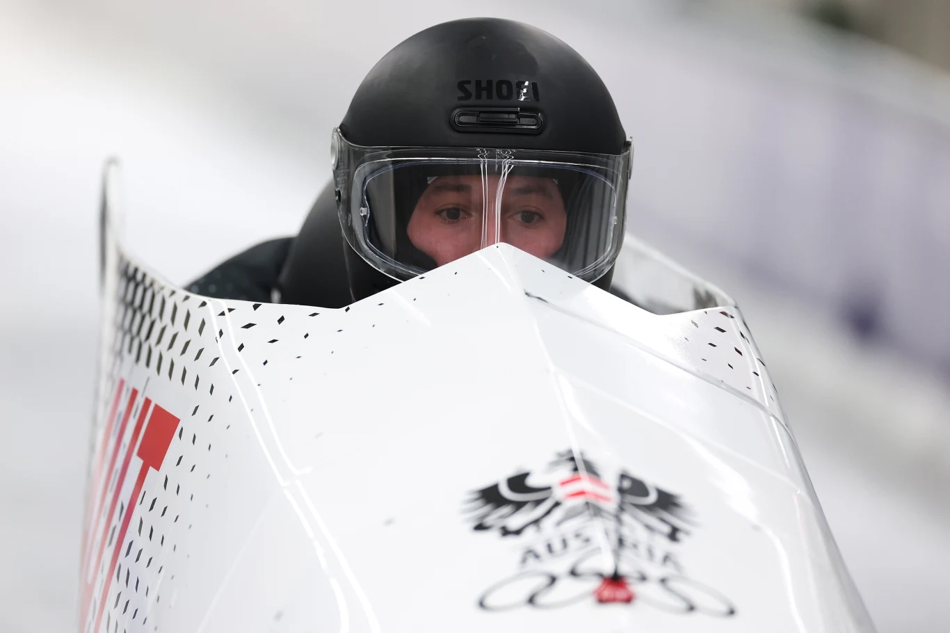 Zawodnik w kasku z przyłbicą prowadzi bobslej z wyraźnym austriackim orłem na przodzie, poruszający się po torze lodowym.