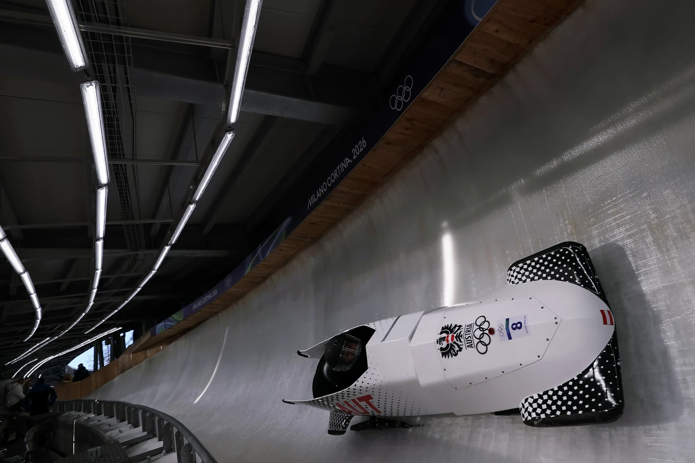 Bobslej z logo austriackim i olimpijskim pędzi po zlodowionym torze pod jasnym oświetleniem hali.