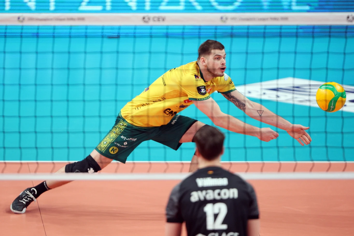 Siatkarz w żółtym stroju wykonuje odbiór piłki dołem podczas meczu siatkówki, w tle widoczny przeciwnik obserwujący akcję.