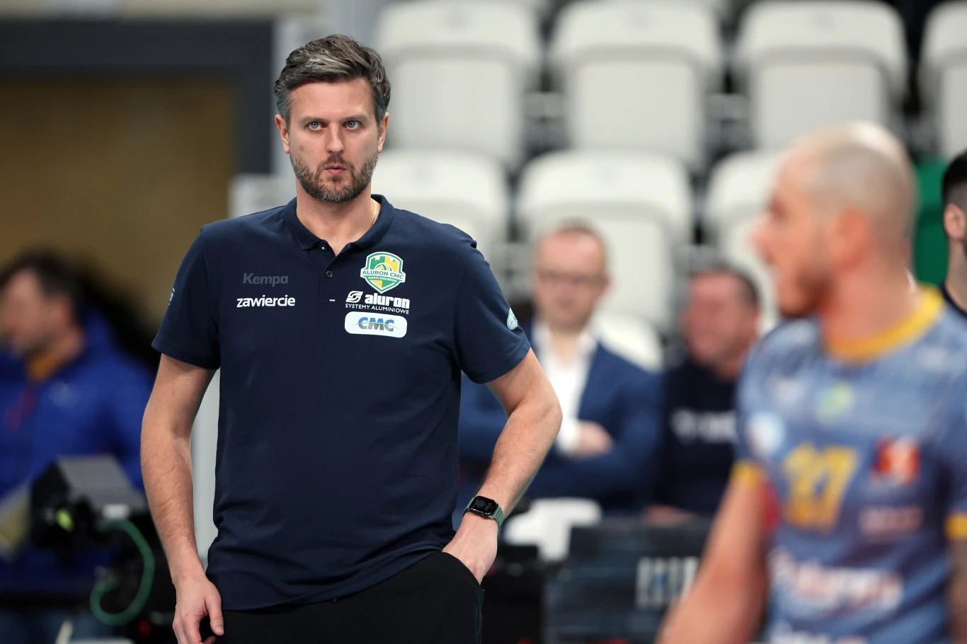 Trener siatkówki w granatowej koszulce z logo klubu obserwuje wydarzenia na boisku, w tle rozmyci widzowie oraz siatkarz w stroju sportowym.