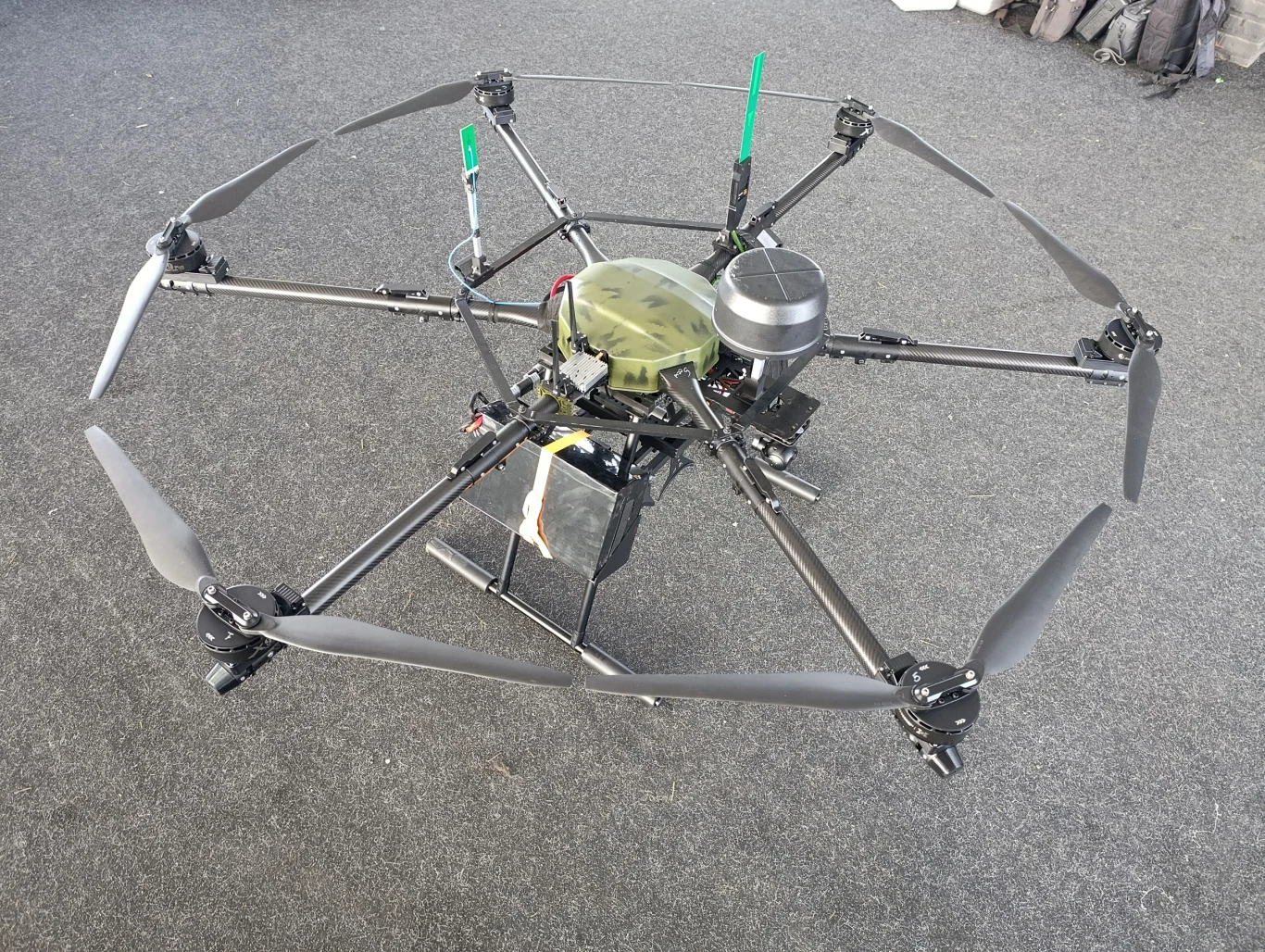 Hexakopter z sześcioma śmigłami o solidnej, czarnej konstrukcji, wyposażony w elektronikę oraz anteny, ustawiony na szarej powierzchni, widoczny z góry.