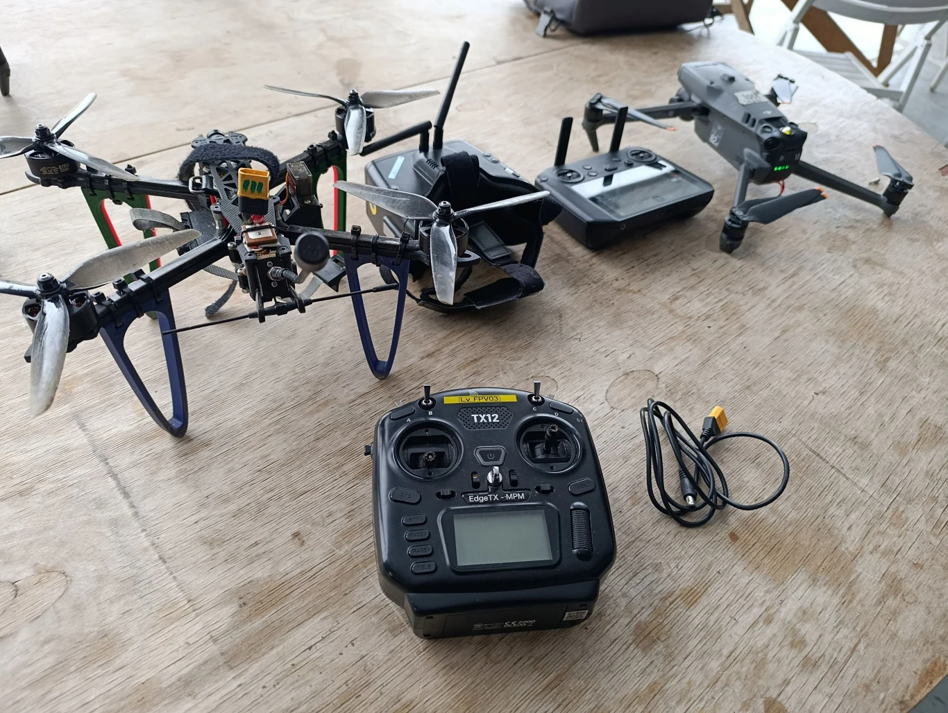 Dwa drony z charakterystycznymi śmigłami i wyeksponowanymi komponentami elektronicznymi ustawione na drewnianym stole. Obok kontroler lotu TX12 oraz przewód do ładowania lub połączenia, na drugim planie widoczny futerał lub torba.