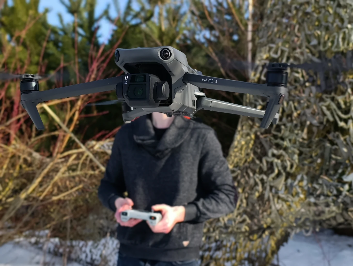 Bezzałogowy dron DJI Mavic 3 unosi się w powietrzu na pierwszym planie, za nim stoi osoba obsługująca urządzenie zdalnie za pomocą kontrolera. W tle widoczna roślinność i śnieg.