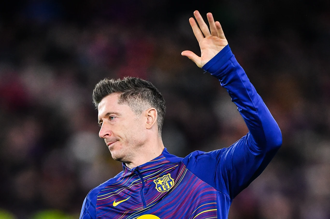 Robert Lewandowski Piłkarz w stroju FC Barcelony unosi dłoń w geście powitania lub pożegnania, na tle rozmytego stadionu.